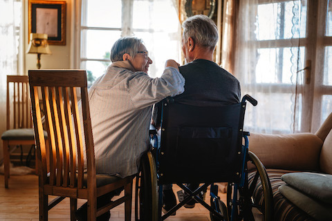 An older lady sits in a wooden chair next to an older man in a wheelchair.